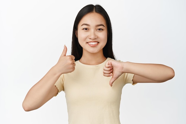 Het portret van een glimlachend aziatisch meisje toont duimen omhoog en duim omlaag, zoals en niet van gebaar dat in een t-shirt op een witte achtergrond staat