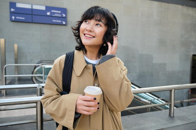 Het portret van een jong studentenmeisje in een koptelefoon drinkt koffiekraampjes op straat met rugzak pendelt t