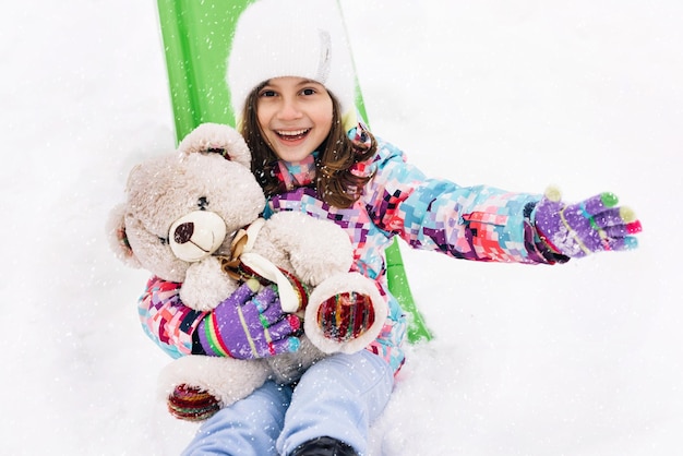 Het portret van een kind met een speelgoedteddybeer zit op een slee en kijkt naar de besneeuwde bergen in de winter