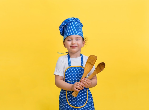 Het portret van een mooi meisje kleedde zich als chef-kok en houdt een houten spatel en een lepel. leuk meisje in een blauwe en gele schort van de chef-kok en glb op een gele ruimte met een exemplaarruimte.