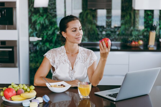 Het portret van een mooie gelukkige vrouw die met laptop werkt tijdens het ontbijt met ontbijtgranen en melk