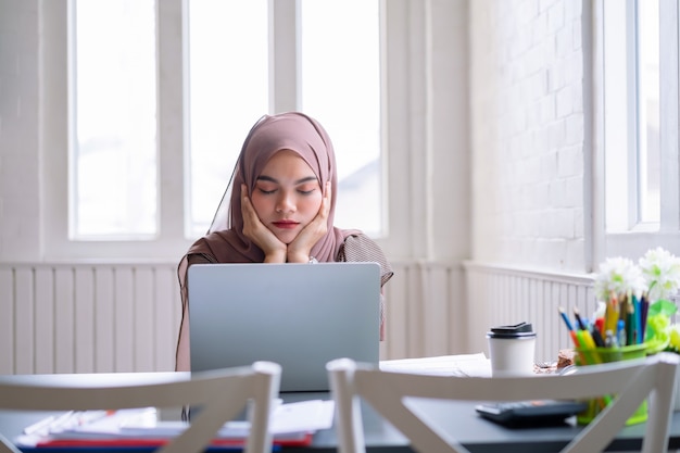 Het portret van een vermoeide moslimvrouwenbeambten die bij de lijst met laptop computer zitten valt na hard het werken in slaap.