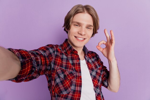 Het portret van een vrolijke, aardige, aantrekkelijke kerel schiet selfie toont een goed teken op een violette achtergrond