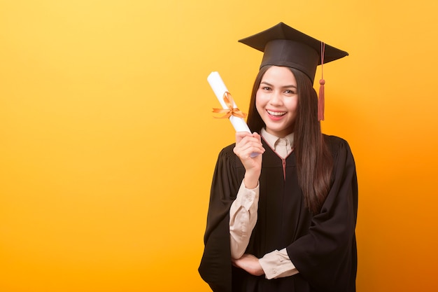 Het portret van gelukkige Mooie vrouw in graduatietoga houdt onderwijscertificaat op gele achtergrond