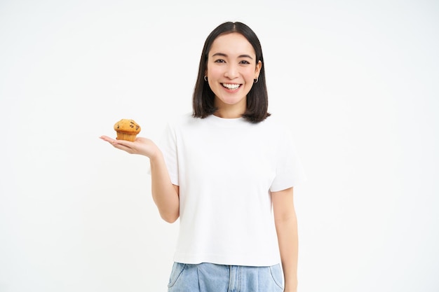 Het portret van glimlachende koreaanse vrouw toont cupcake in haar hand die op witte achtergrond wordt geïsoleerd