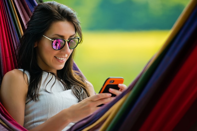 Het portret van jong donkerbruin meisje in zonnebril ligt in een hangmat en speelt met mobiele telefoon. Detailopname.