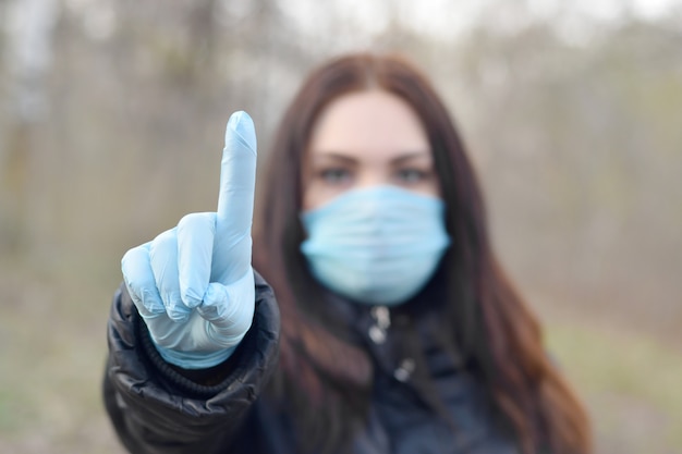 Foto het portret van jonge donkerbruine vrouw in blauw beschermend masker en rubberhandschoenen toont aandachtsgebaar