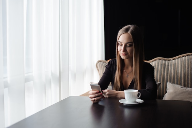 Het portret van jonge onderneemster gebruikt mobiele telefoon terwijl het zitten in comfortabele koffiewinkel tijdens het werkonderbreking.