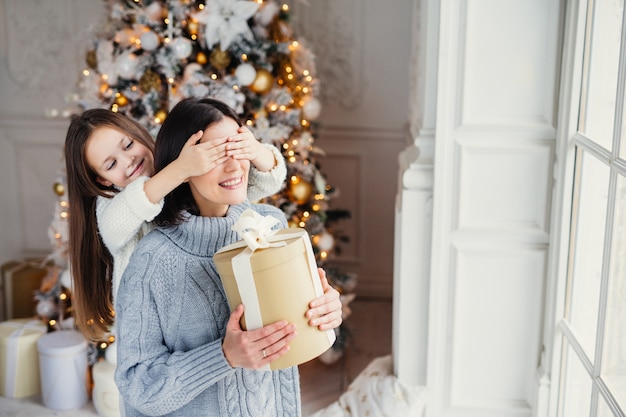 Het portret van meisje sluit de ogen van de moeder, feliciteert haar met nieuwjaar of kerstmis, bevindt zich dichtbij venster in woonkamer, heeft echt wonder en gevoel van vakantie. winter, feest, seizoen