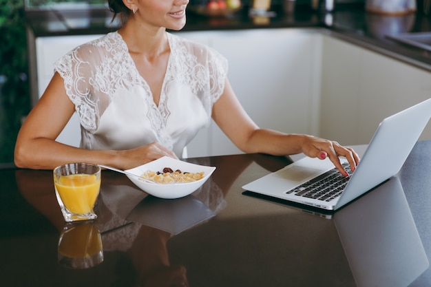 Het portret van mooie jonge vrouw die met laptop werkt tijdens het ontbijt met ontbijtgranen en melk and