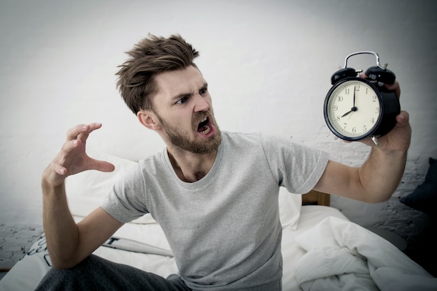 Foto het portret verstoorde de boze jonge mens die bij wekker op slaapkamer gillen