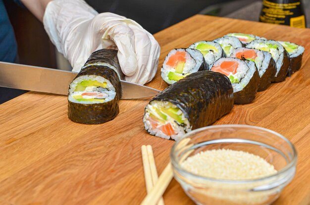 Het proces van het maken van Japanse sushi. Mes in de hand snijdt een close-up van een rol op een houten bord. Vrouwelijke handen in rubberen handschoenen