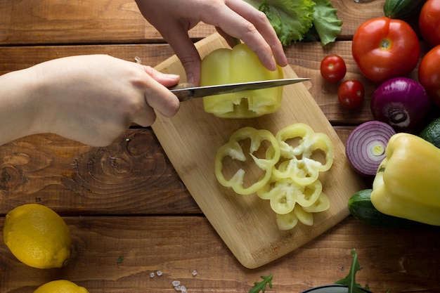 Het proces van het snijden van groenten voor vegetarische salade. Vrouwenhanden snijden tomaten, komkommers, uien, paprika op een houten tafel.