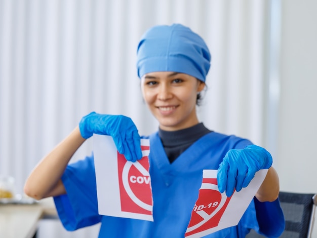 Het rode Covid-19-papieren bord werd verscheurd door een gelukkige mooie arts in een blauw ziekenhuisuniform op een wazige achtergrond toen de pandemie van het coronavirus is geëindigd en het normale leven en de zakelijke veiligheid open zijn.