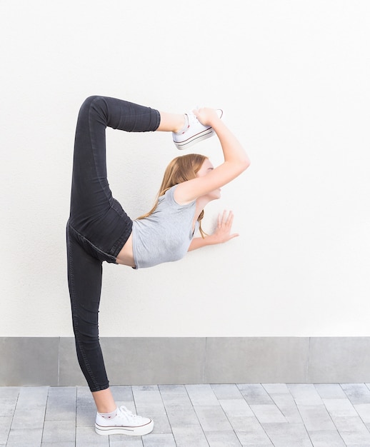 Het roodharige meisje in profiel in ballet stelt haar been boven het hoofd met witte sneakers, zwarte broek en grijs t-shirt op