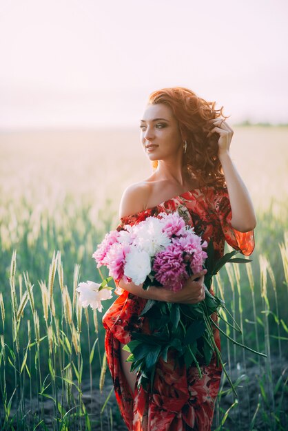 Het roodharige meisje in rode kleding met boeket van pioenen bloeit gebied in de zomer bij zonsondergang. Vrouw geluk.