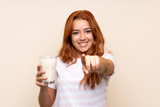 Het roodharigemeisje dat van de tiener een glas melk over geïsoleerde achtergrond houdt wijst vinger op u met een zekere uitdrukking