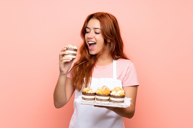 Het roodharigemeisje dat van de tiener veel verschillende minicakes over geïsoleerde roze achtergrond houdt