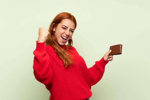 Het roodharigemeisje van de tiener met sweater over geïsoleerde groen houdend een portefeuille