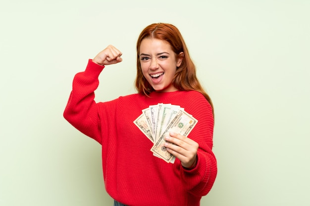 Het roodharigemeisje van de tiener met sweater over geïsoleerde groene achtergrond die heel wat geld neemt