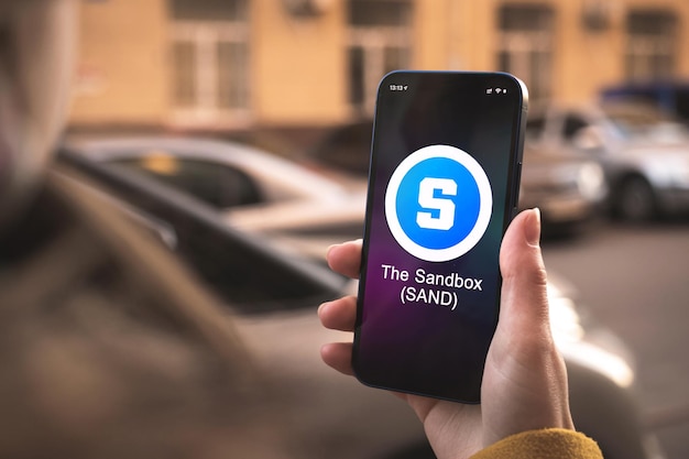 Het Sandbox-cryptocurrency-symbool, SAND-logo. Zakelijk en financieel concept. Hand met smartphone, scherm met crypto icoon close-up