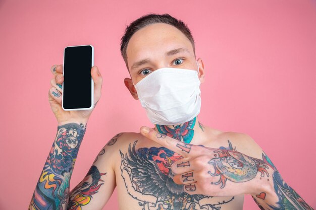 Het scherm van de lege telefoon. portret van een freaky jongeman op roze muur. ongewone look met enorme tatoeages. het dragen van een gezichtsmasker voor bescherming. menselijke emoties, gezichtsuitdrukking, verkoop, advertentieconcept. jeugd.