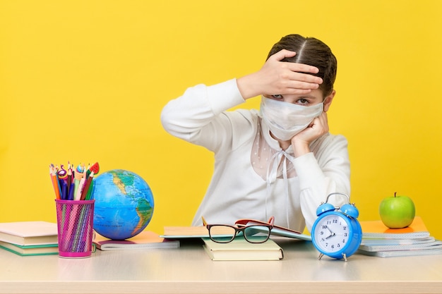 Het schoolmeisje aan haar bureau in een medisch masker op gele achtergrond. Het concept van thuisonderwijs tijdens quarantaine. Terug naar school. Het nieuwe schooljaar. Kinderopvoeding concept.