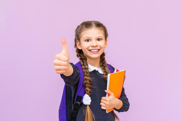 Het schoolmeisje geeft een duim omhoog en glimlacht breed Een klein meisje met een rugzak en notitieboekjes om op een roze geïsoleerde achtergrond te studeren