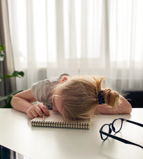 Het schoolmeisje was het thuisonderwijs beu en viel in slaap aan de tafel op een notitieboekje met een potlood in haar hand. Afstandsonderwijs tijdens het coronavirus.