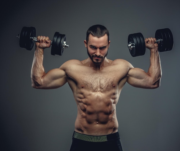 Het shirtless gespierde mannetje houdt halters met armen omhoog op een grijze achtergrond.