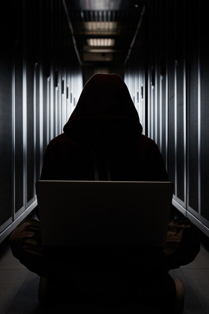 Het silhouet van de hacker in een kap de inbreker zit met de computer tussen servers in het donker