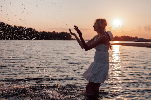 Het silhouet van een slanke vrouw bij zonsondergang een mooie jonge blonde in een zomerjurk en een stro h...