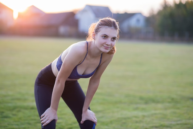 Het slanke blondemeisje speelt sporten en voert yoga uit stelt in de zomer gras behandeld stadion op een zonsondergangachtergrond. vrouw die oefeningen op de yogamat doet.