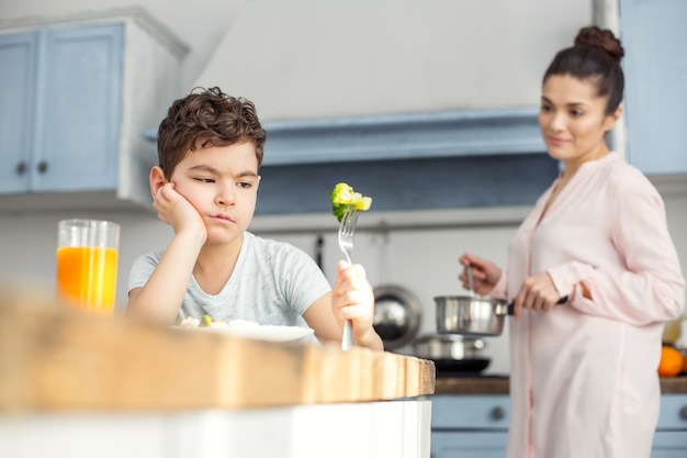 Het smaakt slecht. aantrekkelijke ongelukkige donkerharige jongetje gezond ontbijten en kijken naar de groene groente op zijn vork en zijn moeder staande in de