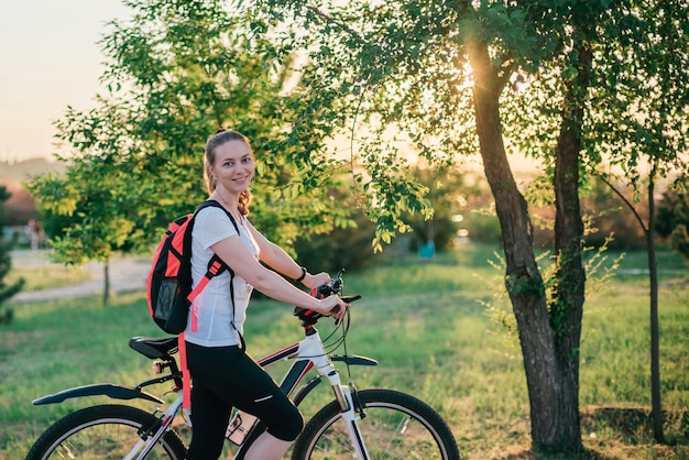 Het sportieve jonge blondemeisje in sportkleding met een rugzak berijdt een fiets en glimlacht naar de camera