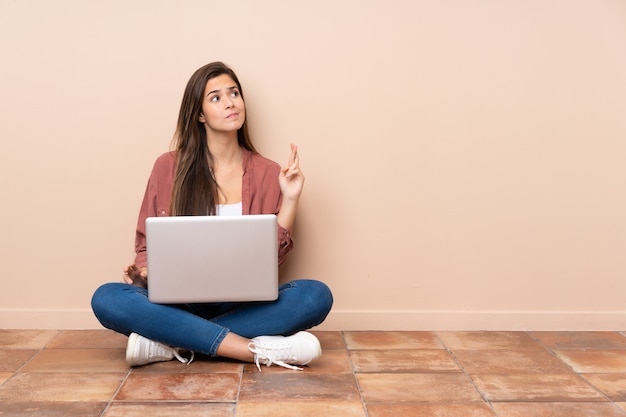 Het studentenmeisje van de tiener zitting op de vloer met laptop met vingers die en het beste kruisen wensen