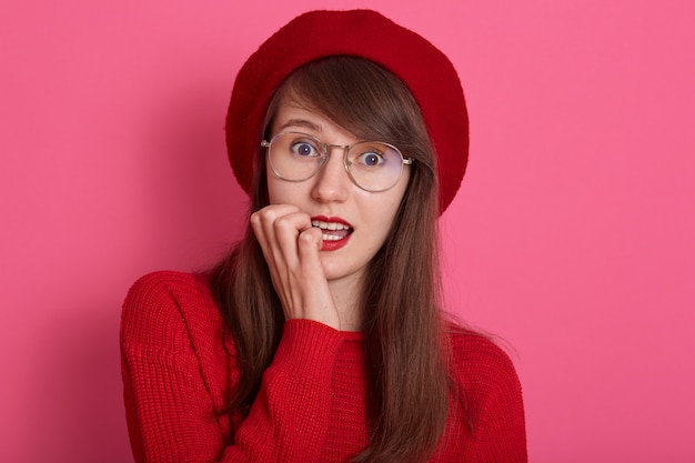 Het studioportret van mooie jonge Kaukasische vrouw die haar vinger bijten, kijkt bang, heeft perfecte huid en donker haar, heldere pommade maakt omhoog, dragend sweater en baret