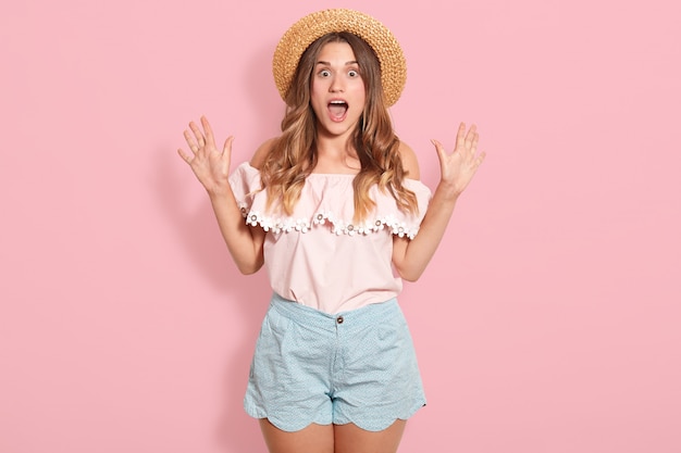 Het studioschot van jonge vrouw die roze de zomerblouse, blauwe korte broek, zonnebril en de zomerhoed dragen die zich met open mond in schok bevinden, kijkt verrast, uitdrukkend verbazing. Mensen en emoties concept.