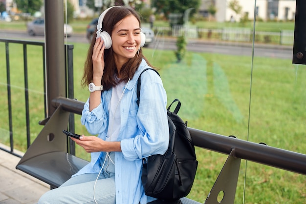 Het tienermeisje luistert naar de muziek door witte hoofdtelefoons in een openbaar vervoerpost terwijl zij op tram wacht.