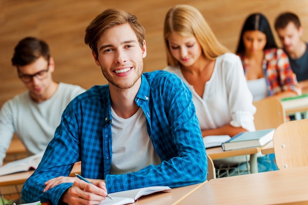 Het tonen van toewijding aan het onderwijs. knappe jongeman die in notitieblok schrijft en naar de camera kijkt terwijl hij aan de receptie in de klas zit