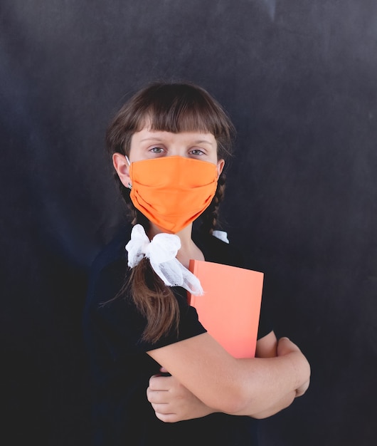 Het trieste meisje in een oranje masker op een zwarte achtergrond met een notitieboekje terug naar school