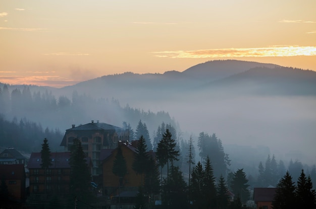 Het vakantiedorp herbergt gebouwen op achtergrond van mistige blauwe bergheuvels die met dicht nevelig net bos onder heldere roze hemel bij zonsopgang worden behandeld. Berglandschap bij dageraad.