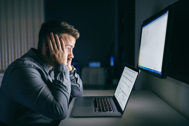 Het vermoeide jonge Kaukasische hoofd van de werknemersholding in handen en het bekijken computermonitor terwijl het zitten laat op nacht in het bureau.