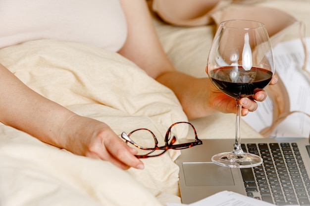 Het volwassen vrouw ontspannen in bed met een glas rode wijn en de laptop computer