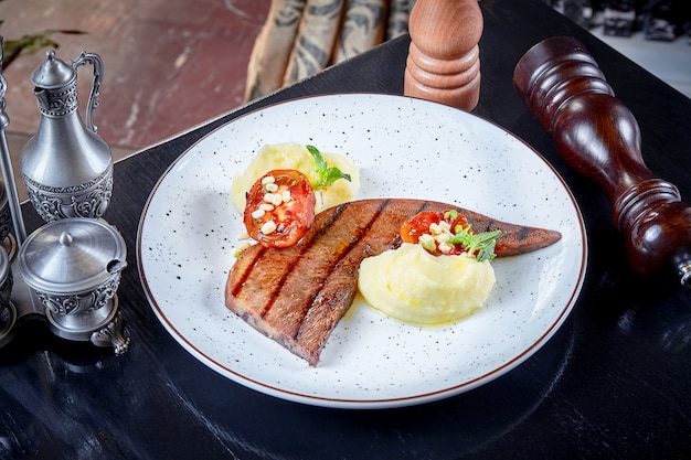 Het vooraanzicht over het rundvleeslapje vlees met aardappelpuree en een geroosterde tomaat diende op witte plaat. Premium Japanse rundertong. Japanse Wagyu Steak. Russische keuken. BBQ-maaltijd. Voedsel achtergrond