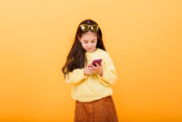 Het vrij glimlachende meisje houdt smartphone. Leuk kind op gele achtergrond, modern kinderenconcept.