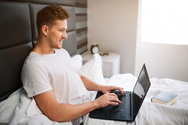 Het vrolijke aardige jonge mensenwerk in bed vroege moring. guy houdt laptop vast en typt op toetsenbord. glimlach en positief. daglicht.