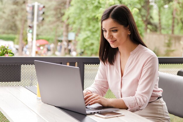 Het vrolijke jonge onderneemster glimlachen die met energie aan computer aan terras in koffie werken