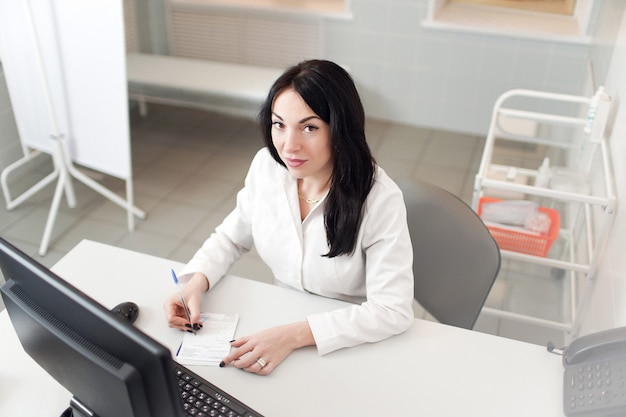 Het werken van de vrouwelijke Arts aan laptop computer, het schrijven voorschriftblocnote met verslaginformatie over bureau in het ziekenhuis of kliniek, Gezondheidszorg en medisch concept