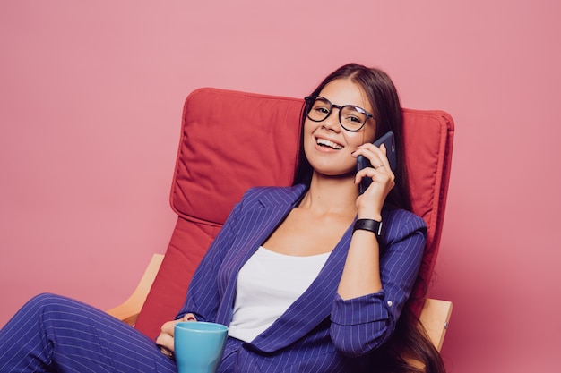 Het zekere brunette met blije uitdrukking in gestreept donkerblauw kostuum, zittend in rode stoel, houdend kop van koffie, heeft een aantrekkelijke blik, sprekend op de telefoon, over roze achtergrond.